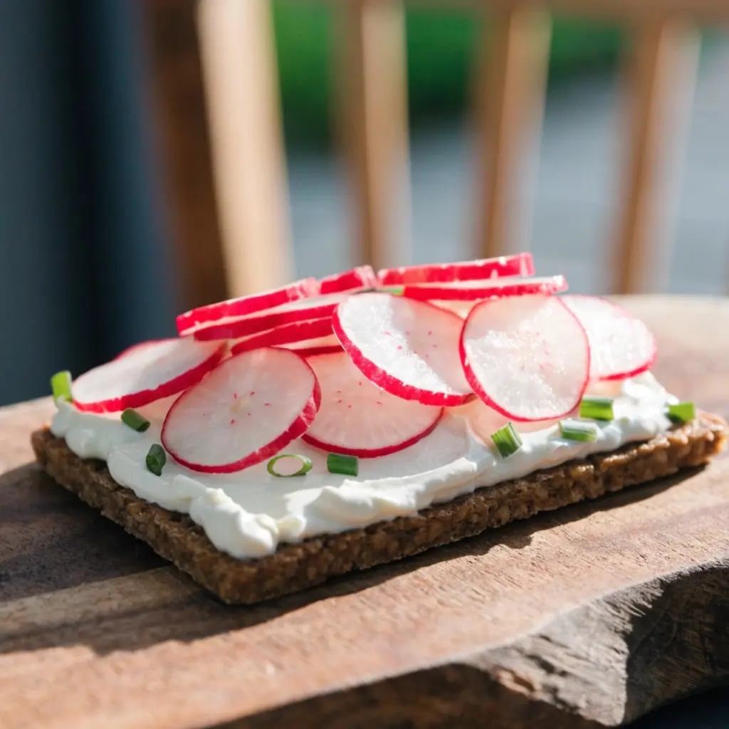 Vollkorn-Knäckebrot mit Frischkäse, dünn geschnittenen Radieschen und frischem Schnittlauch auf einem Holzbrett angerichtet
